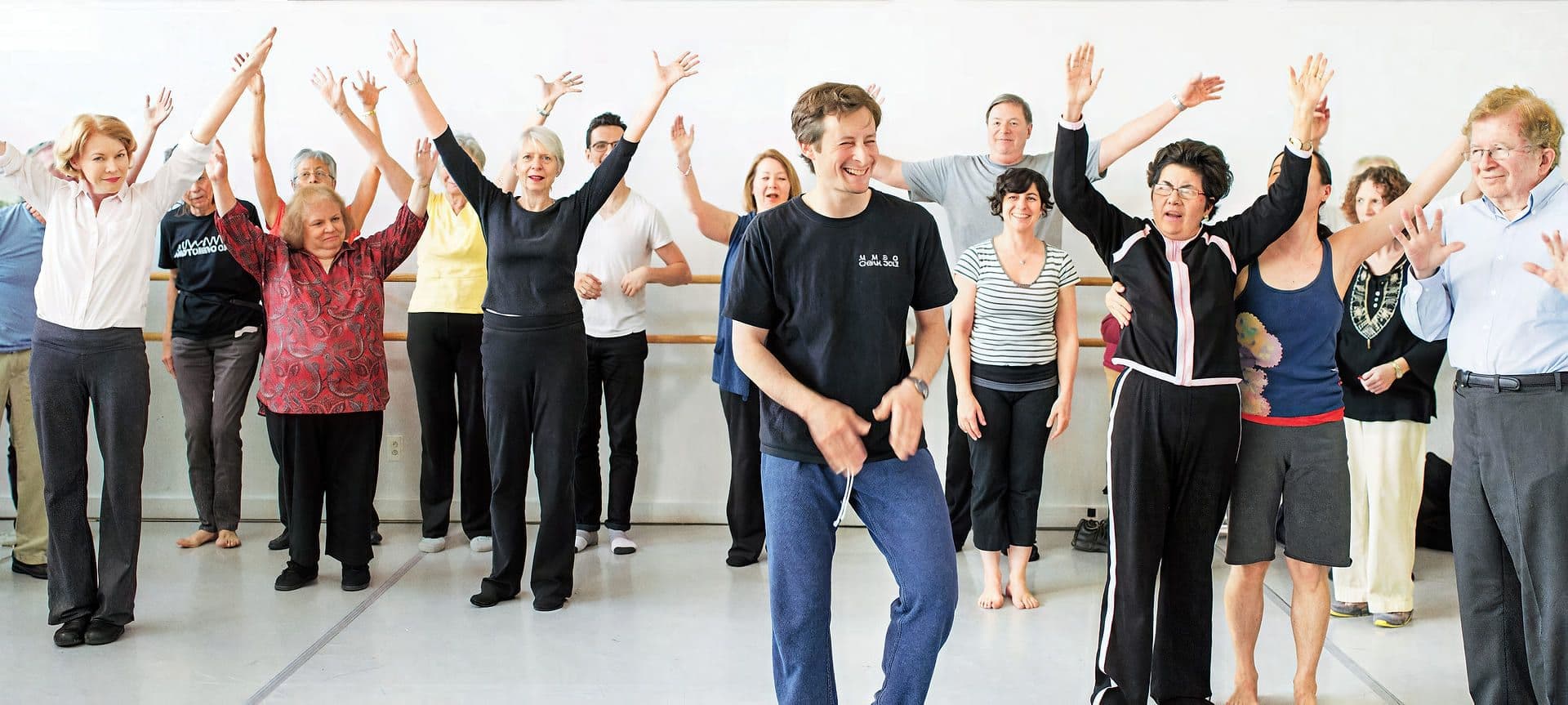 Dance for PD: a dance class for people with Parkinson’s People with Parkinson’s dancing with raised arms in a bright studio, dance teacher David Leventhal in front